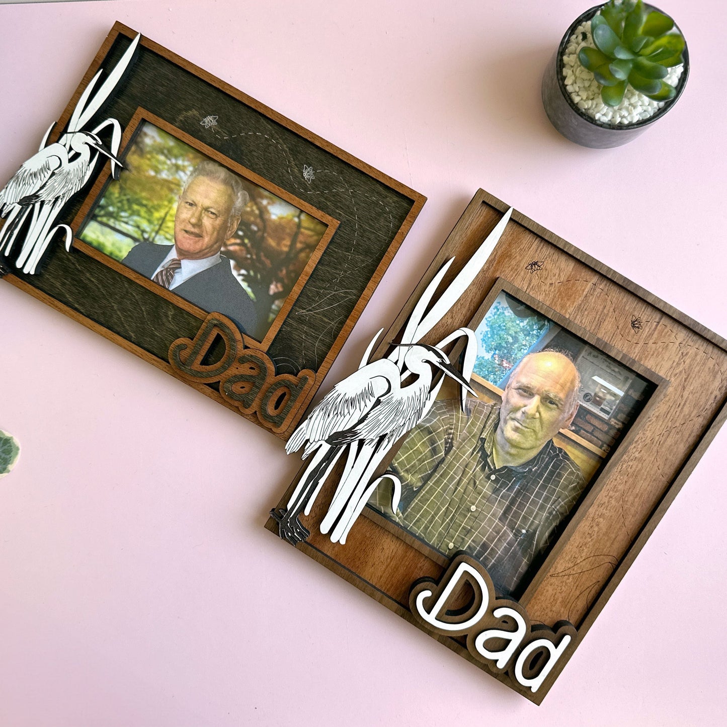 Two wooden photo frames with 'Dad' engraved, each containing a photo of an elderly man, on a light pink surface with plants.