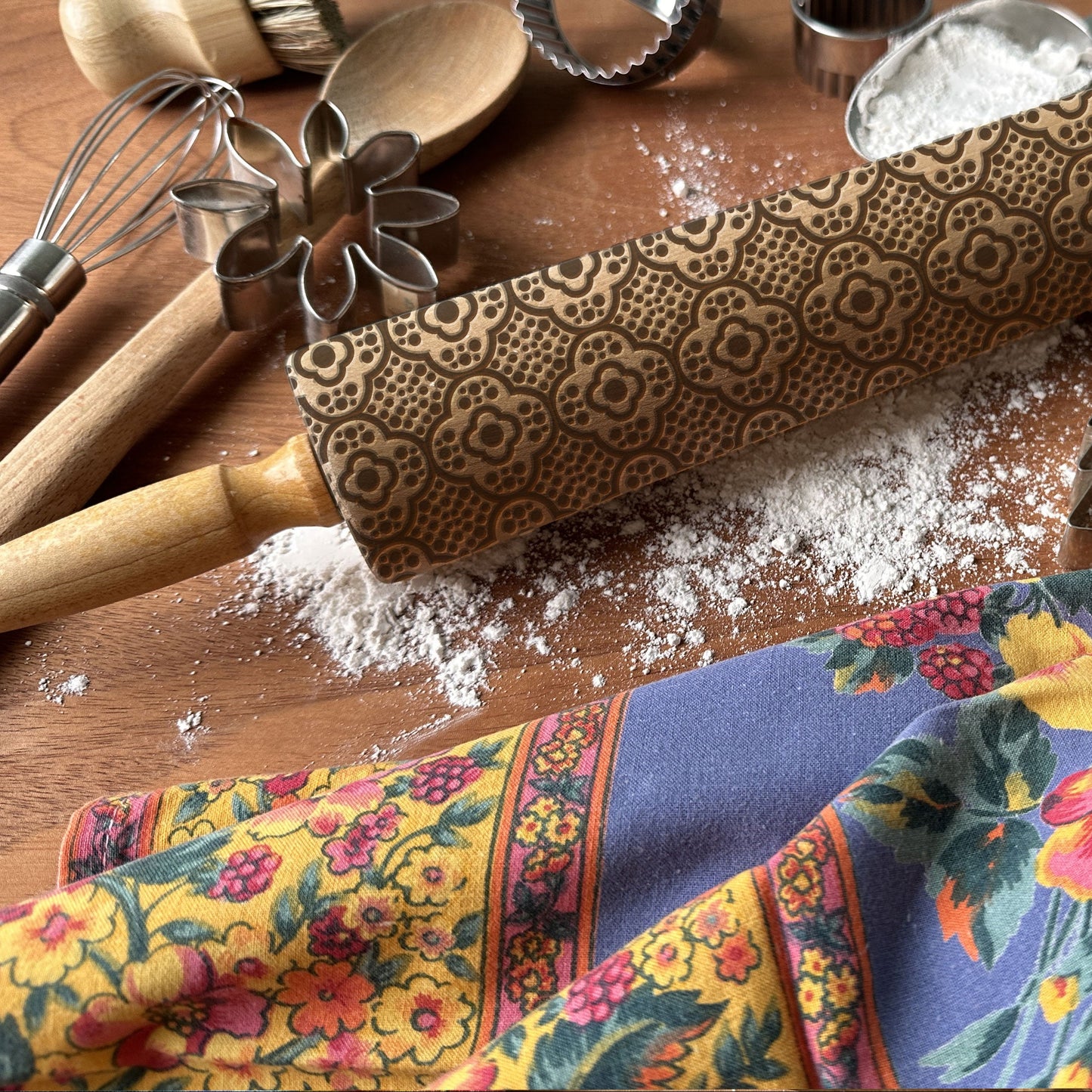 Wooden rolling pin with floral pattern on a wooden surface with baking tools and a colorful towel.