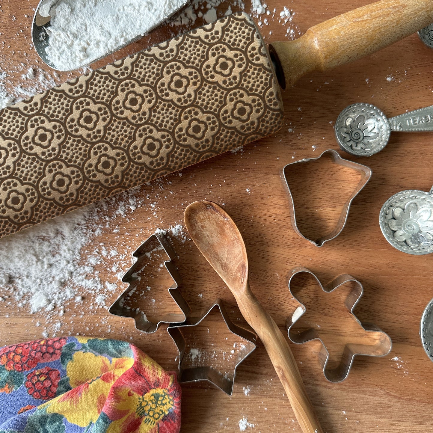 Baking tools including a patterned rolling pin, cookie cutters, and measuring spoons on a wooden surface with flour.