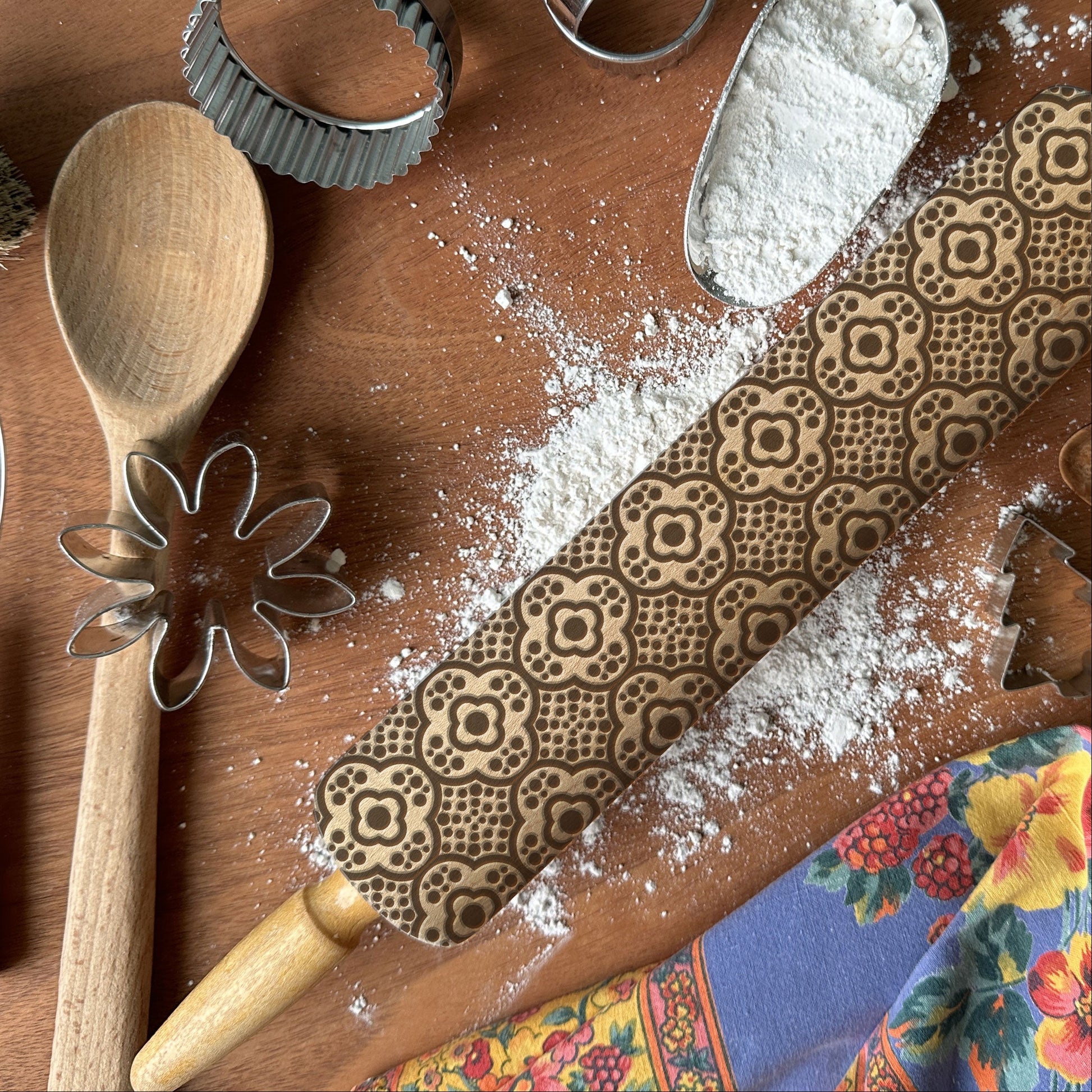 Baking tools including a wooden spoon, rolling pin with decorative patterns, and flour on a wooden surface.