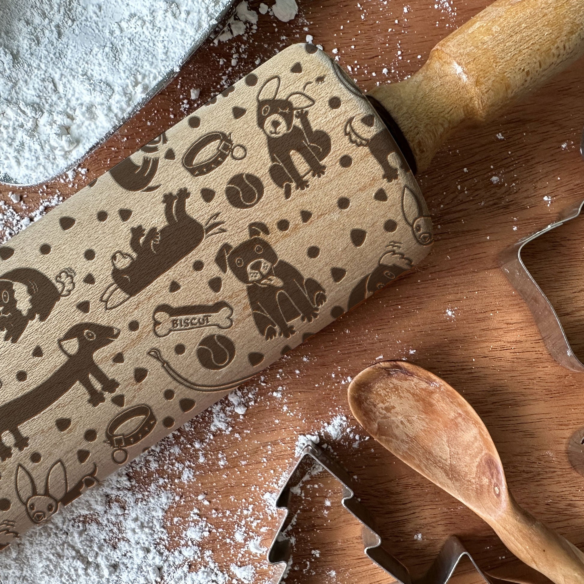 Wooden rolling pin with dog patterns on a wooden surface with flour and cookie cutters.
