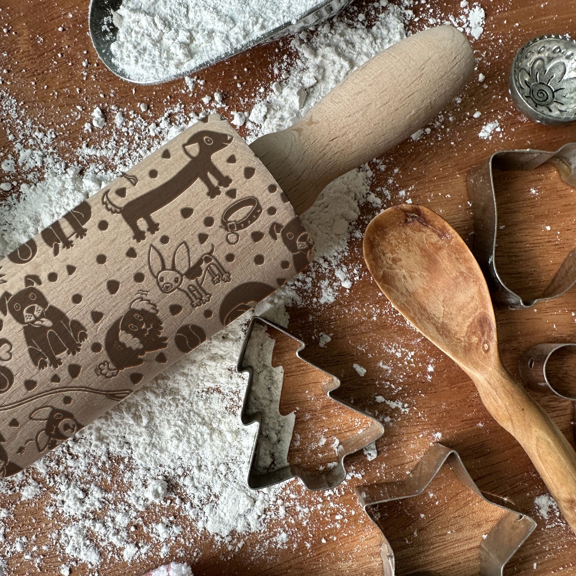 Rolling pin with embossed designs on a flour-dusted surface with cookie cutters and a wooden spoon.