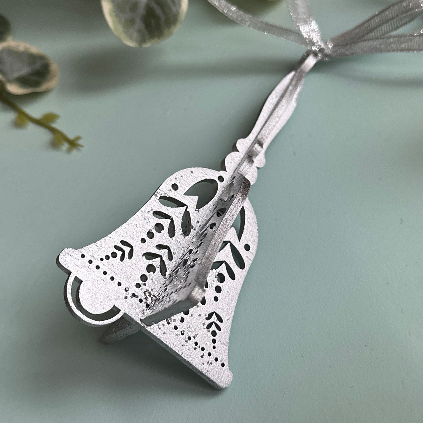 Silver bell-shaped pendant on a light blue background with green leaves.