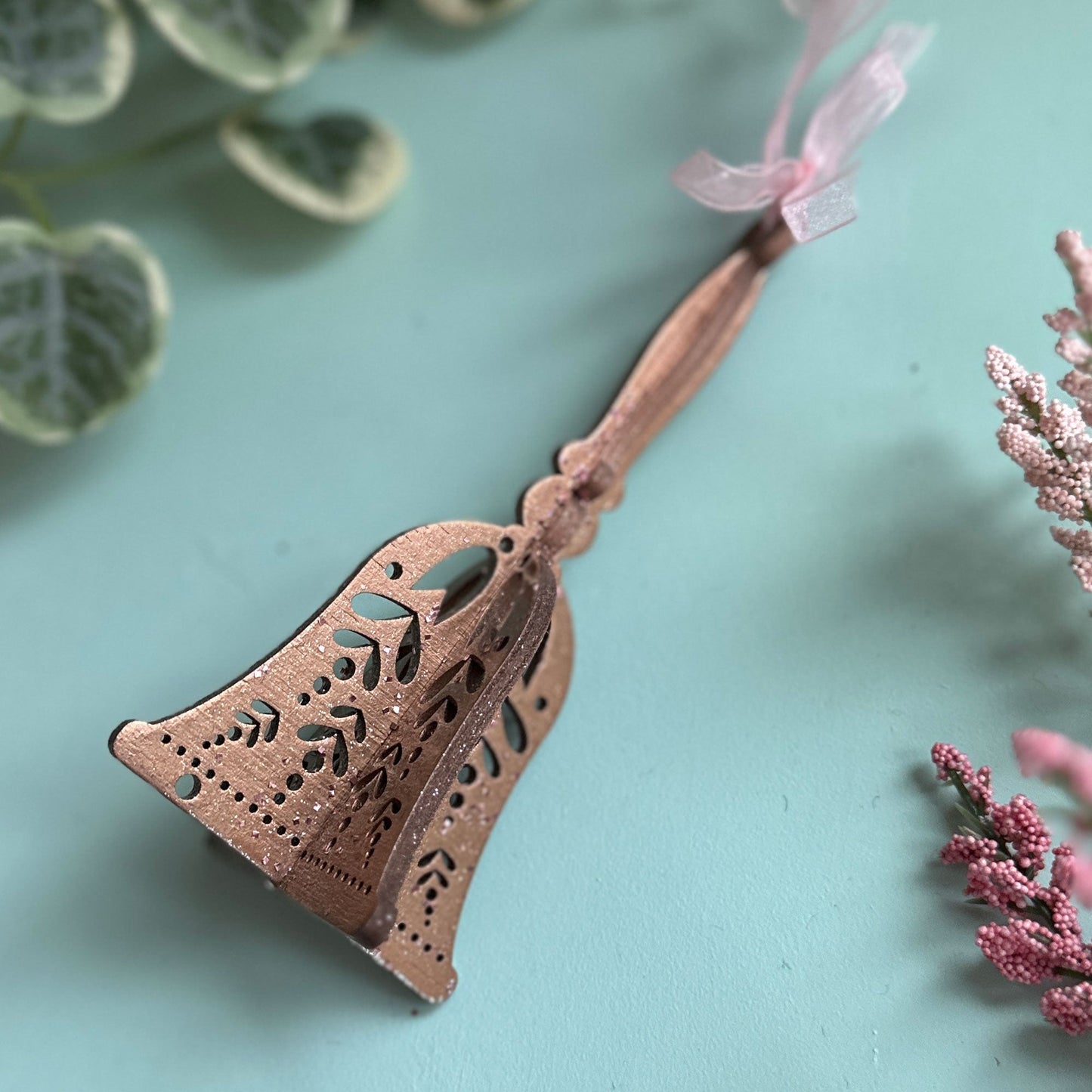 Decorative metal bell with a pink ribbon on a light blue background with green leaves and pink flowers.