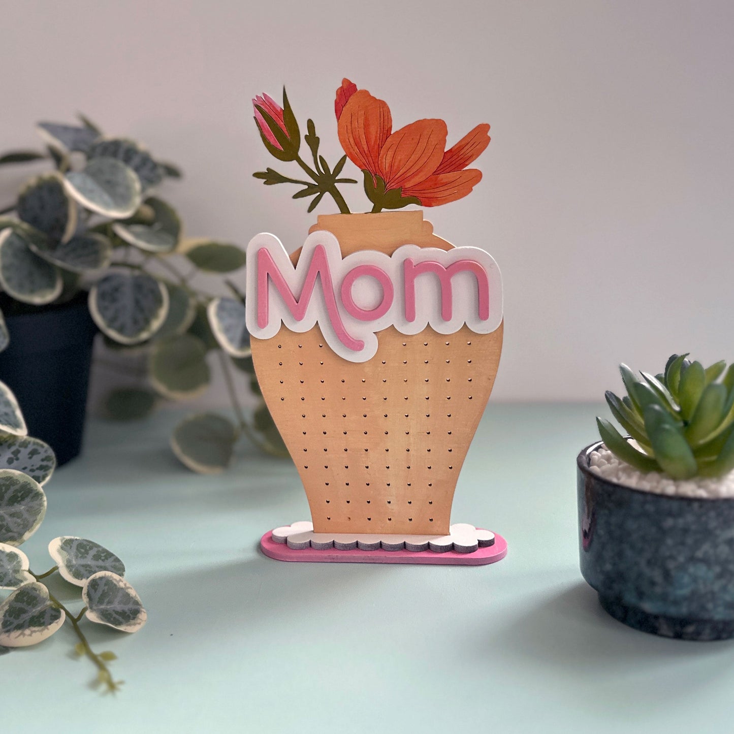 Decorative earring stand with 'Mom' in a vase, surrounded by plants on a light surface.