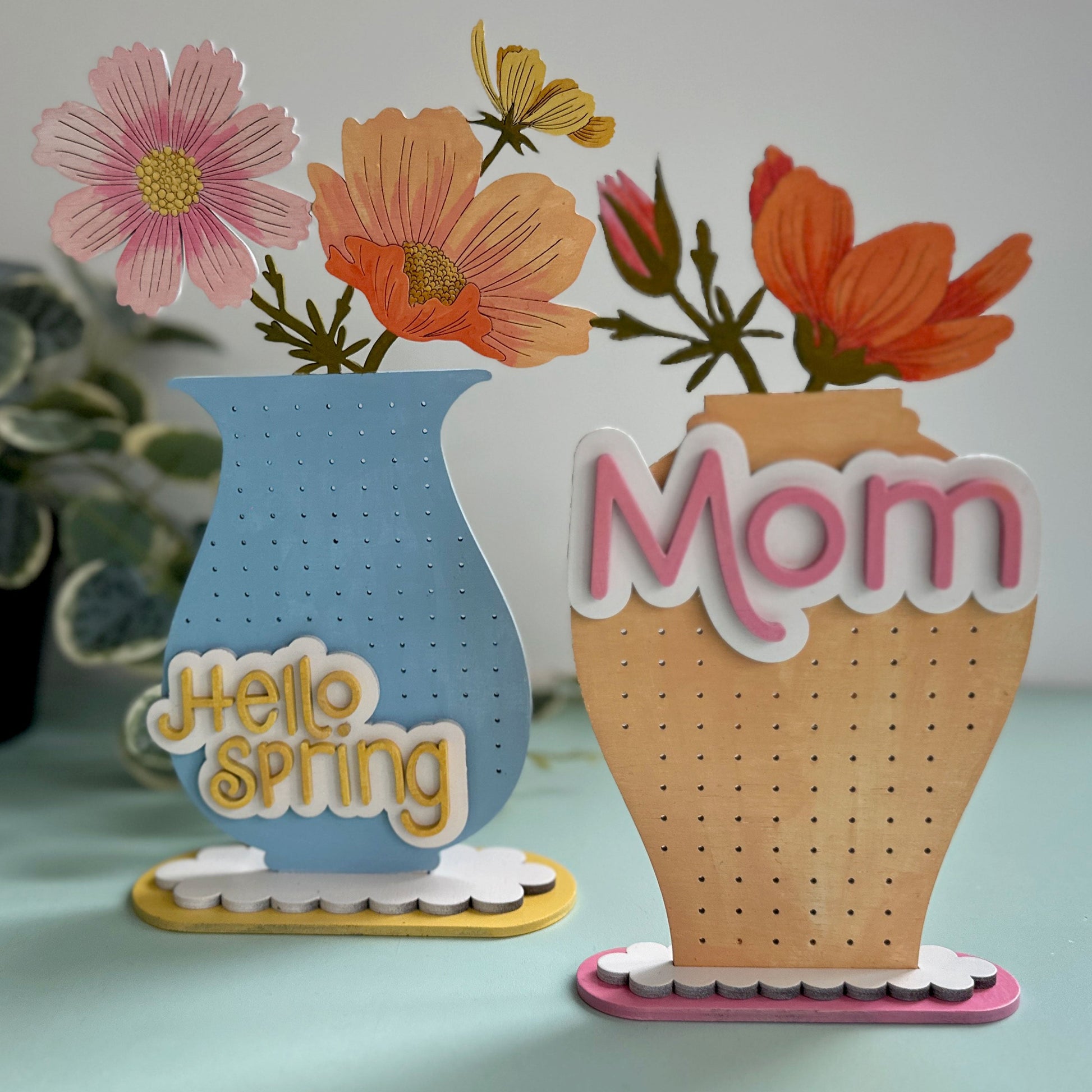 Two earring stands with decorative vases with flowers and 'Mom' text on a light surface.