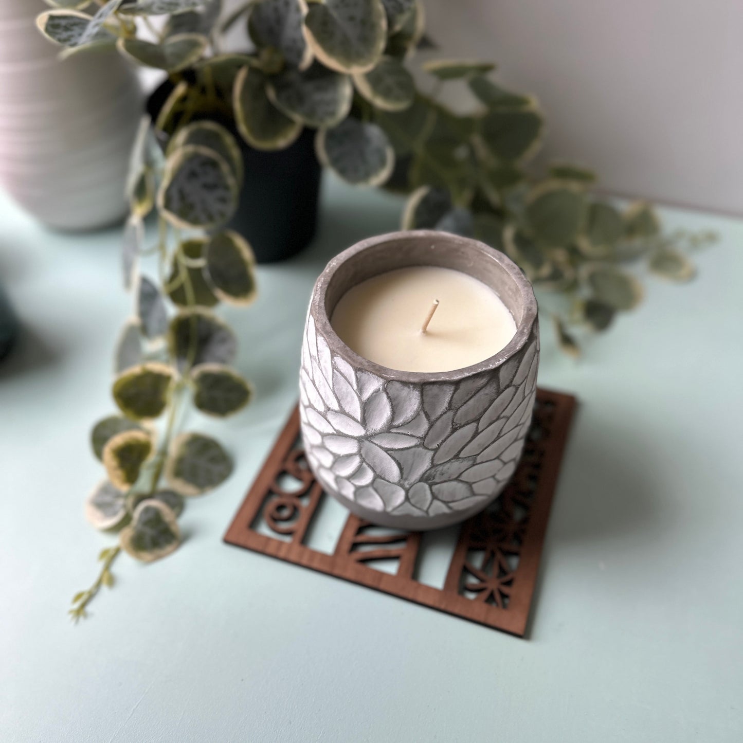 Decorative candle in a leaf-patterned holder on a trivet with a plant in the background