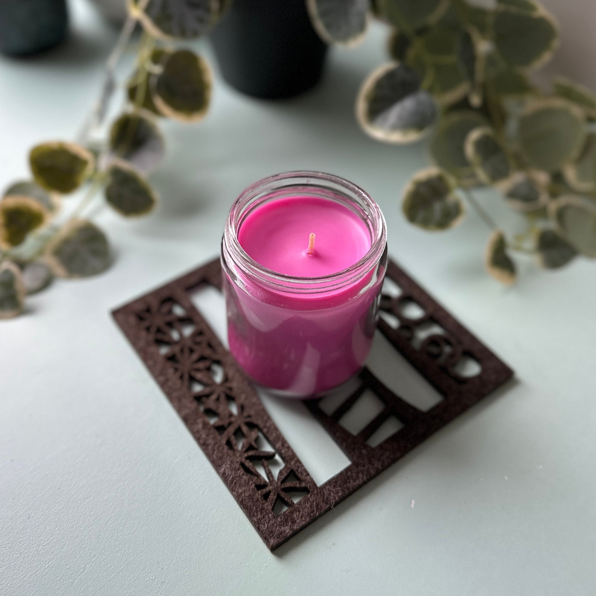 Pink candle in a glass jar on a decorative trivet with plants in the background