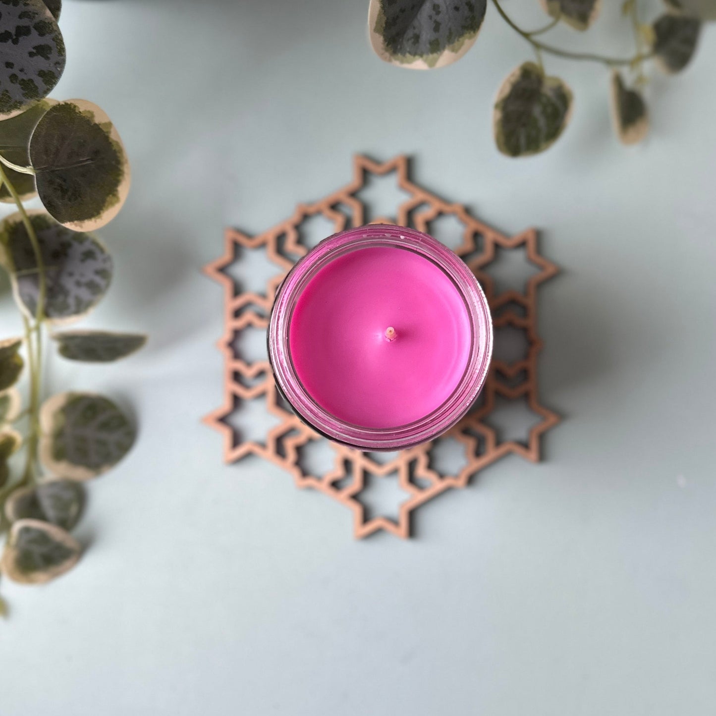 Pink candle on a decorative trivet on a white surface with green leaves in the background