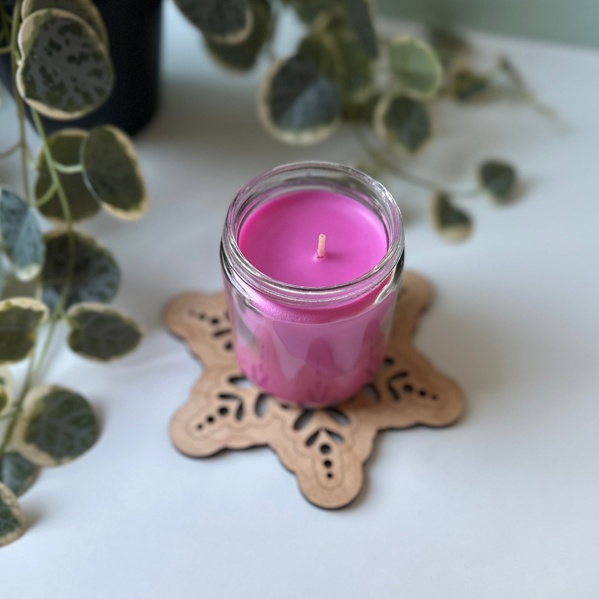 Pink candle in a glass jar on a decorative trivet with green leaves in the background