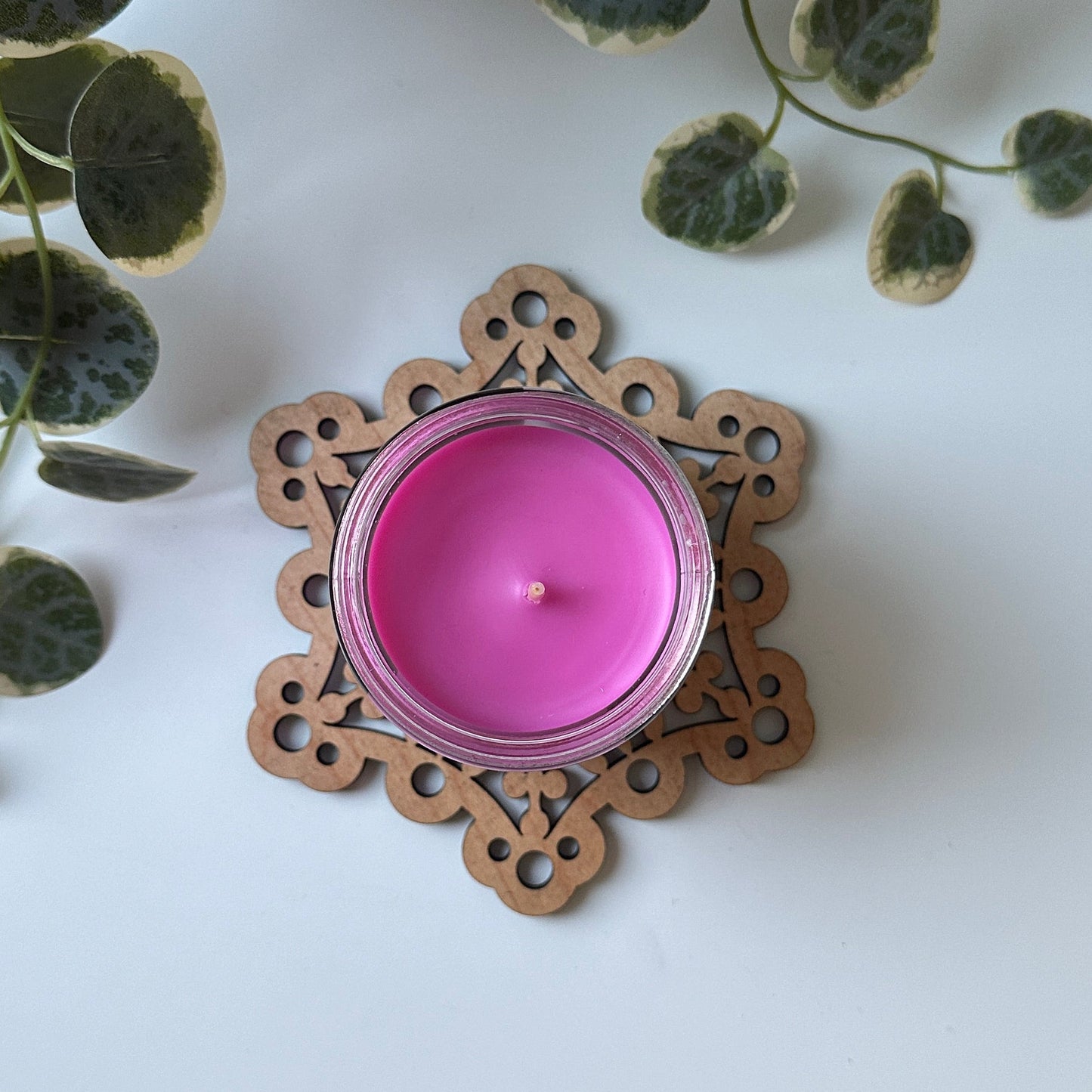 Pink candle on a decorative trivet on a white surface with plants around