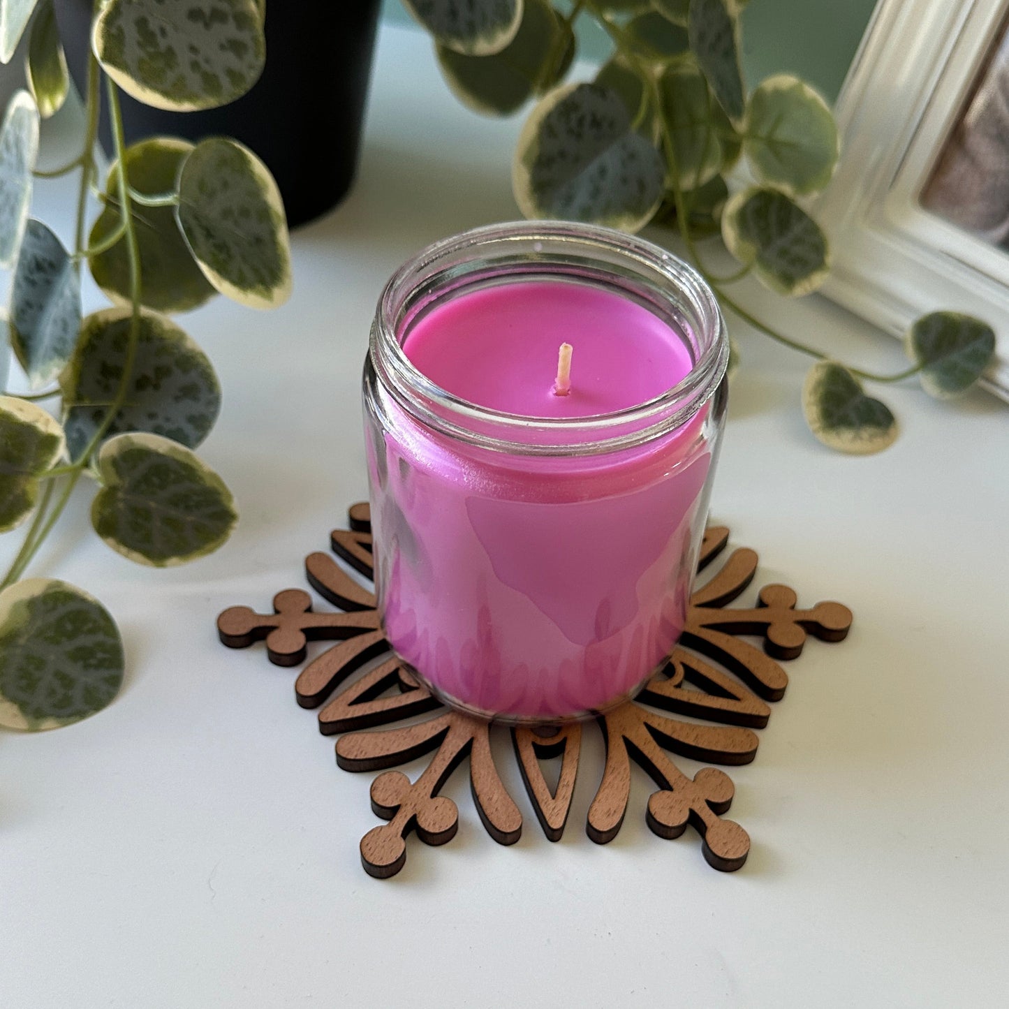 Pink candle in a glass trivet on a decorative coaster with a plant and photo frame in the background.