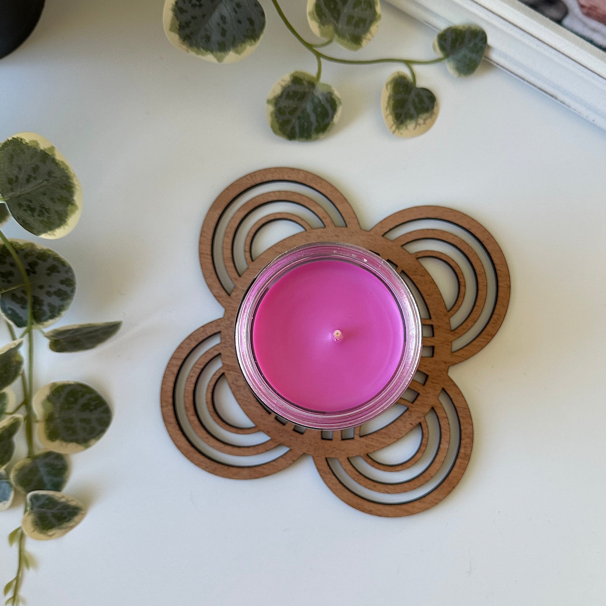 Pink candle in a decorative wooden trivet on a white surface with green leaves and a photo frame.