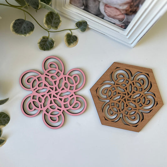 Pink and wooden geometric coasters on a white surface with green leaves.