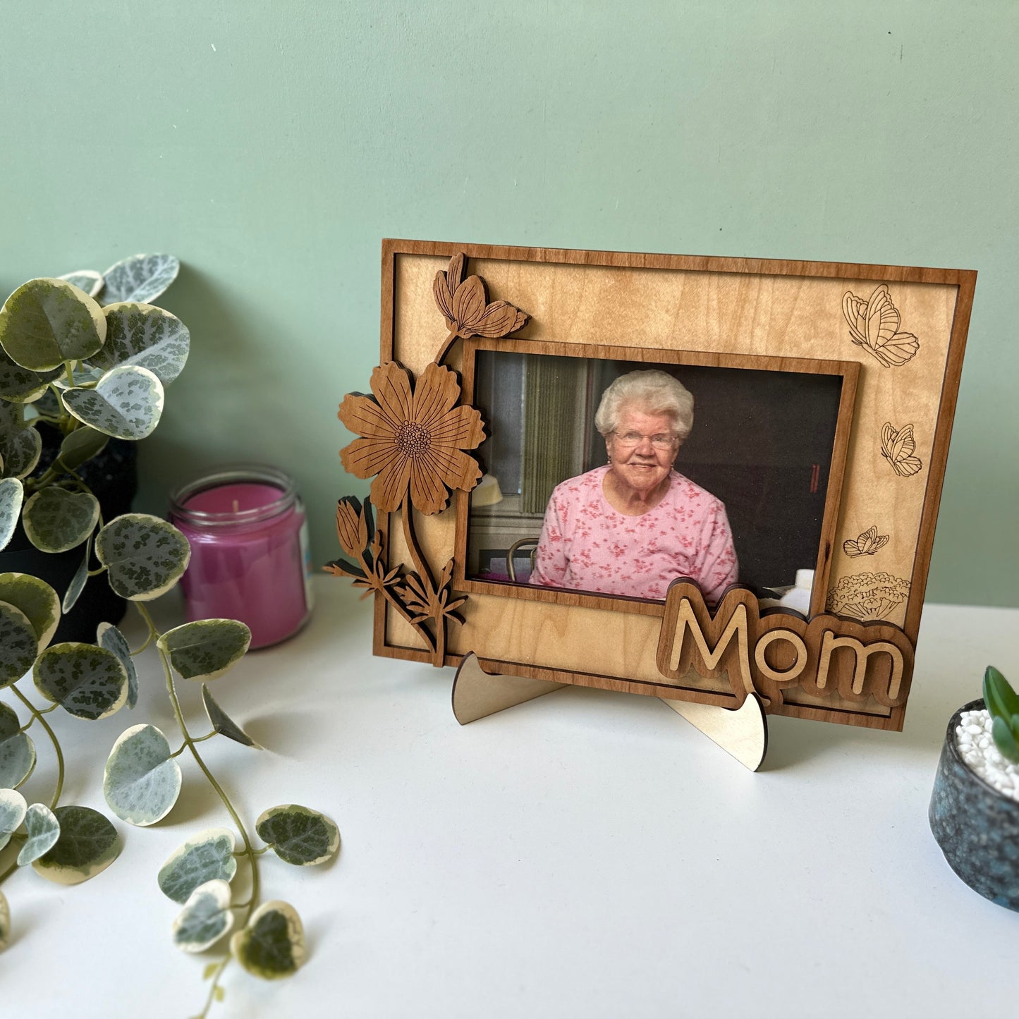 Wooden photo frame with a picture of an elderly woman, labeled 'Mom', on a surface with plants and a candle.