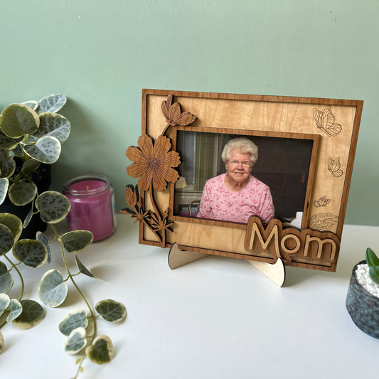 Wooden photo frame with a picture of an elderly woman, labeled 'Mom', on a surface with plants and a candle.