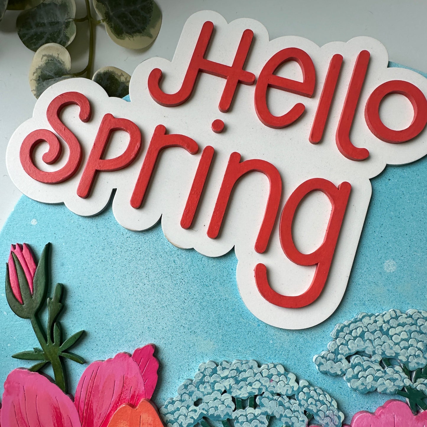 Decorative sign with 'Hello Spring' text surrounded by colorful flowers on a light blue background