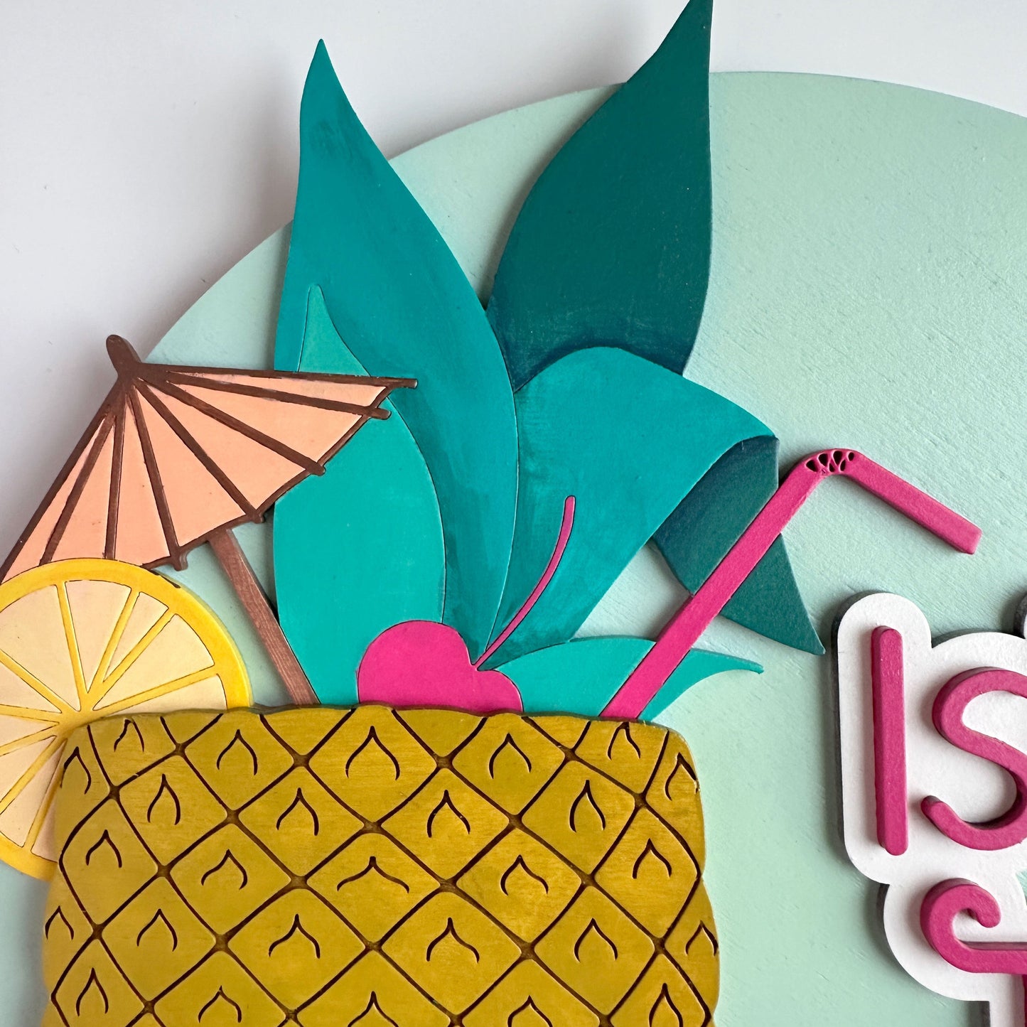 Decorative paper craft of a pineapple with a pink straw, lemon slice, and umbrella on a light green background.