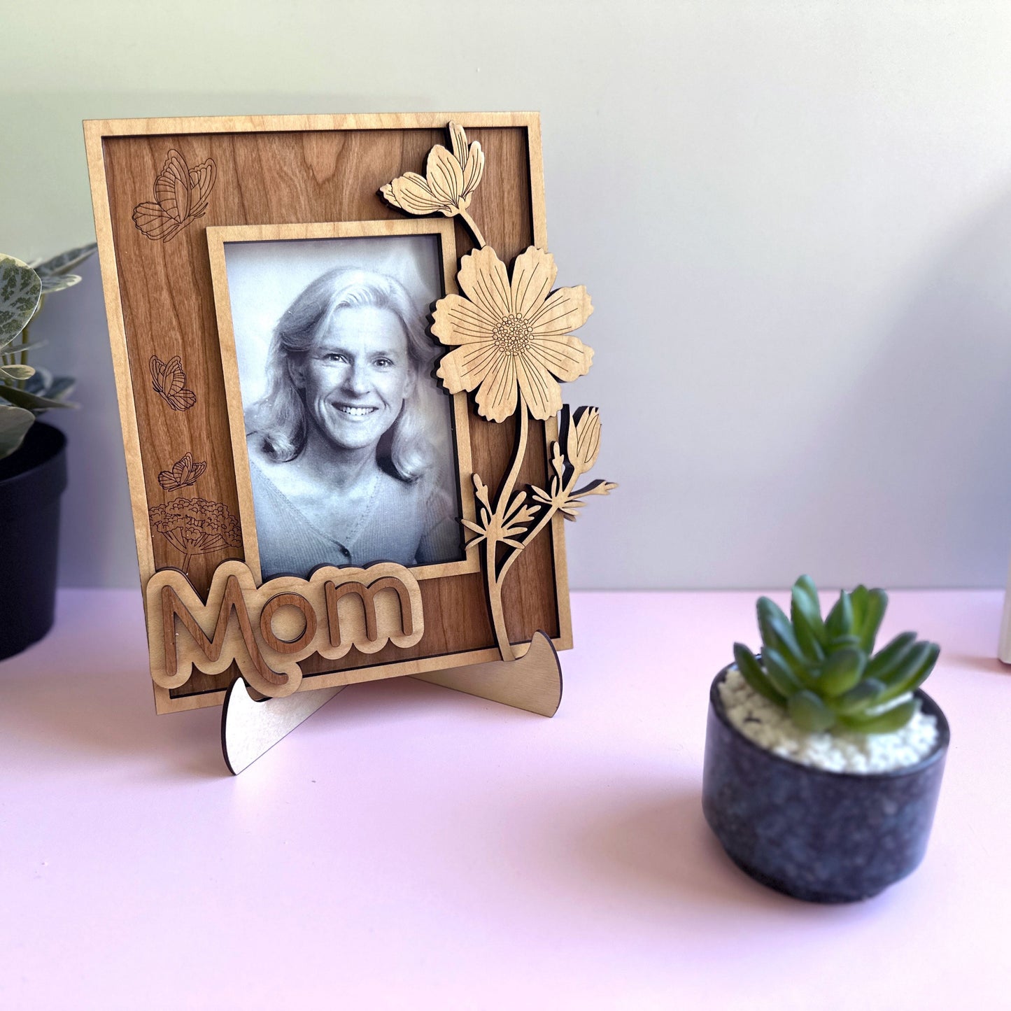 Wooden photo frame with 'Mom' engraving holding a black and white photo of a woman, placed on a surface with plants.