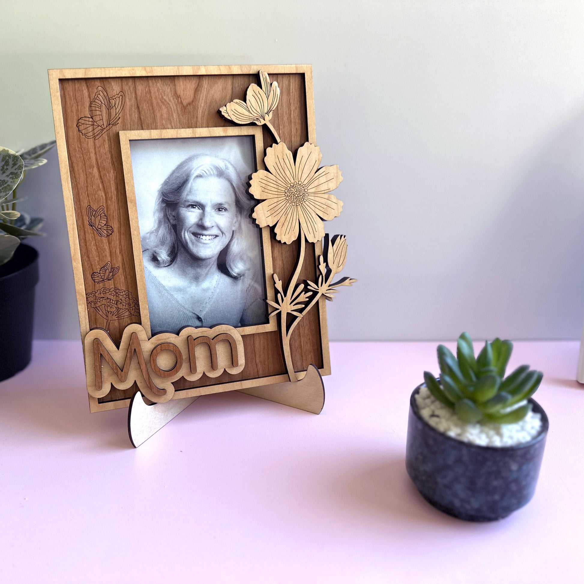 Wooden photo frame with 'Mom' engraving holding a black and white photo of a woman, placed on a surface with plants.