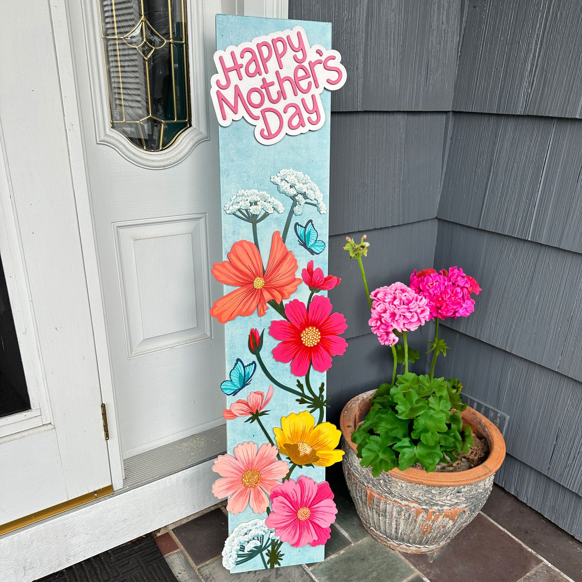 Decorative sign with flowers and 'Happy Mother's Day' text next to a potted plant on a porch.