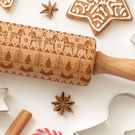 Wooden embossing rolling pin with Christmas designs on a white surface with cookies and spices.