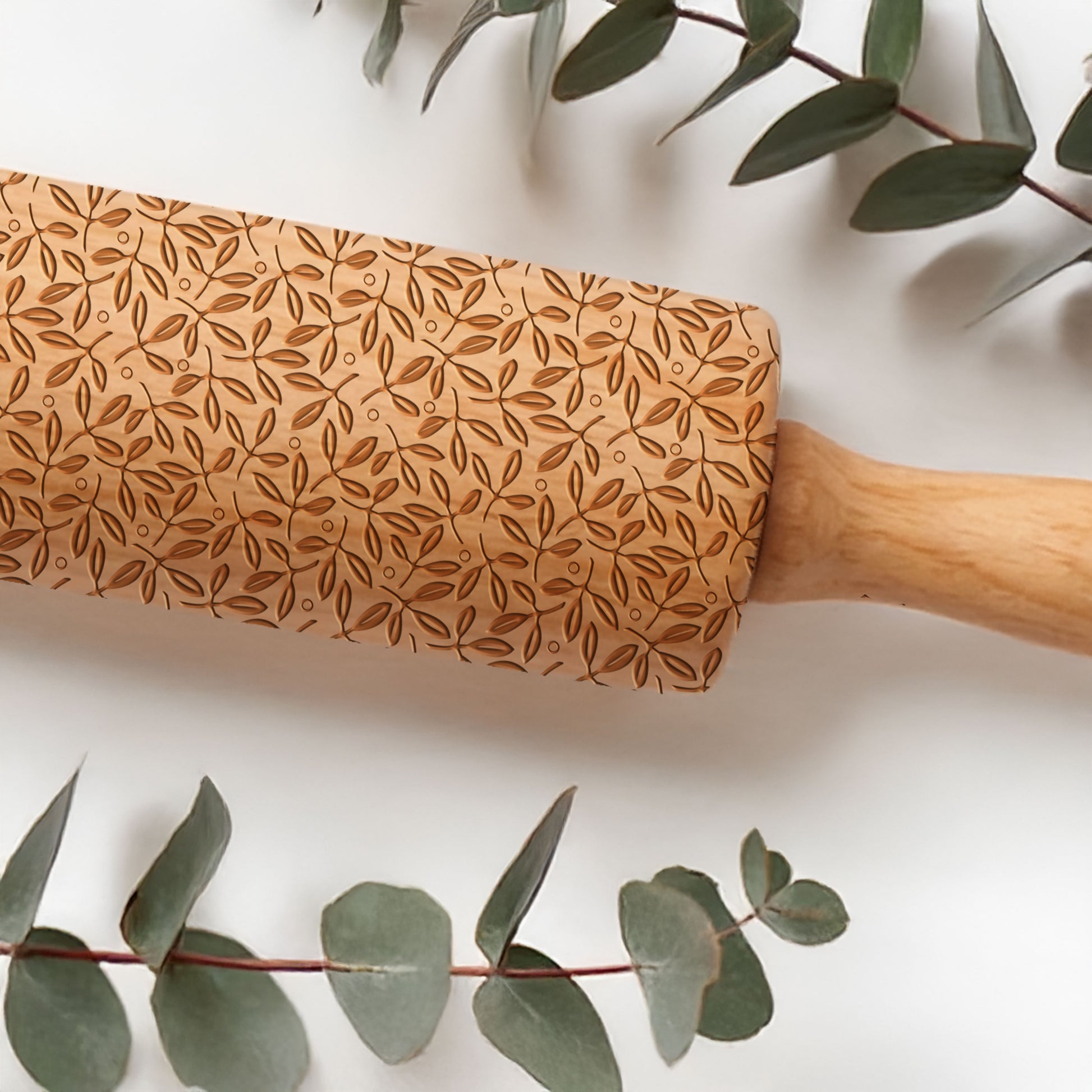 Wooden rolling pin with leaf pattern on a white background with eucalyptus leaves.