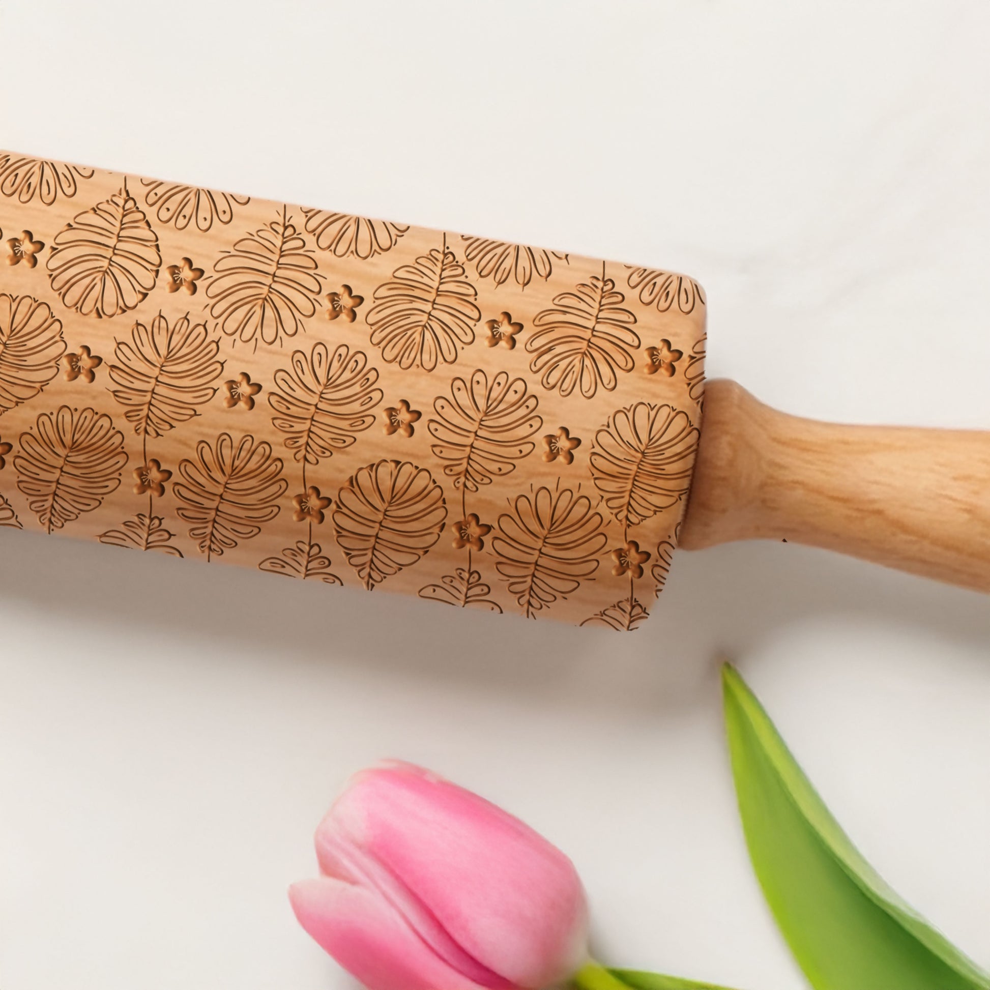 Wooden rolling pin with leaf patterns on a white background with a pink tulip and green leaf.