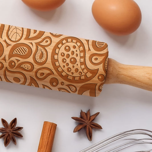 Wooden rolling pin with intricate patterns on a white surface with eggs, star anise, cinnamon stick, and whisk.