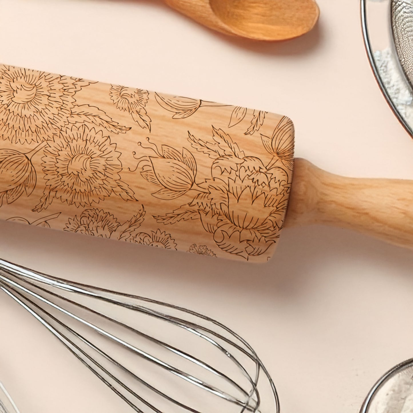 Wooden rolling pin with floral patterns on a light pink surface with kitchen utensils.
