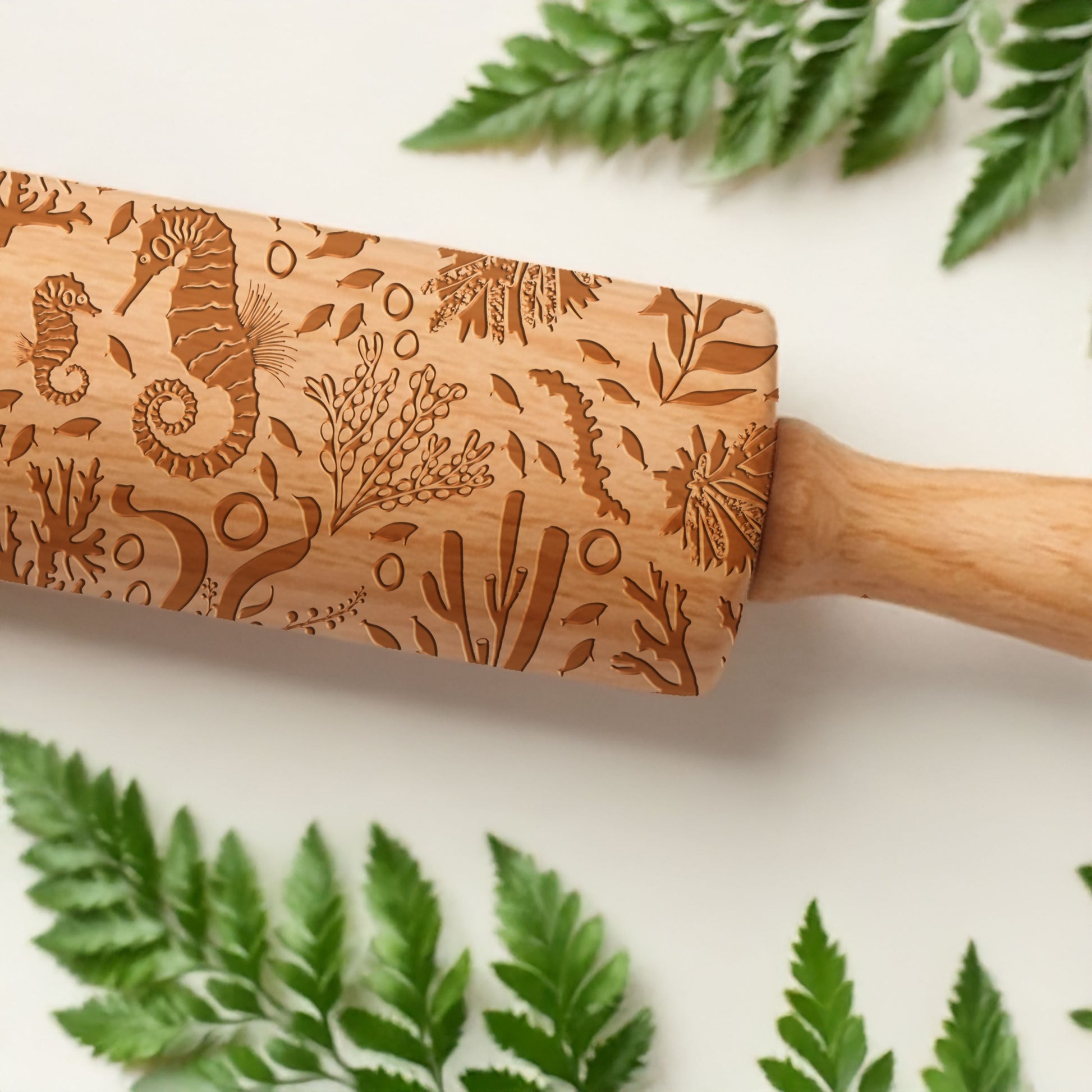 Wooden rolling pin with engraved seahorse and plant designs on a white background with green leaves.