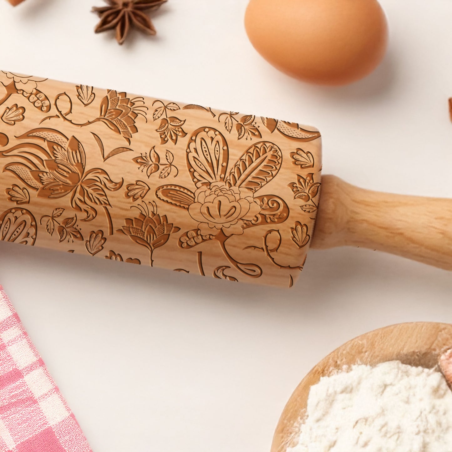 Wooden embossing rolling pin with floral and butterfly patterns on a kitchen counter with flour and eggs.