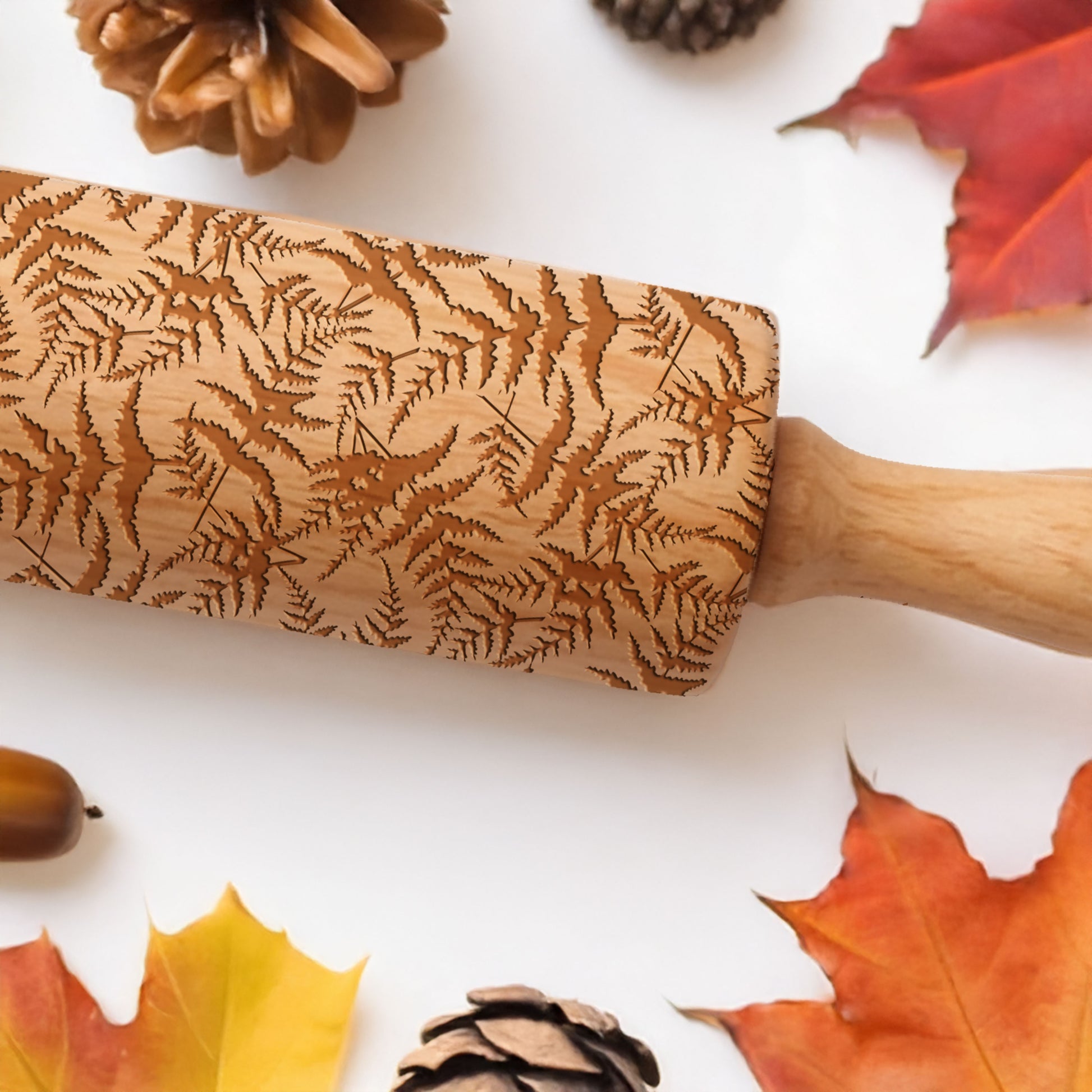 Wooden rolling pin with leaf pattern surrounded by autumn leaves and pinecones on a white background