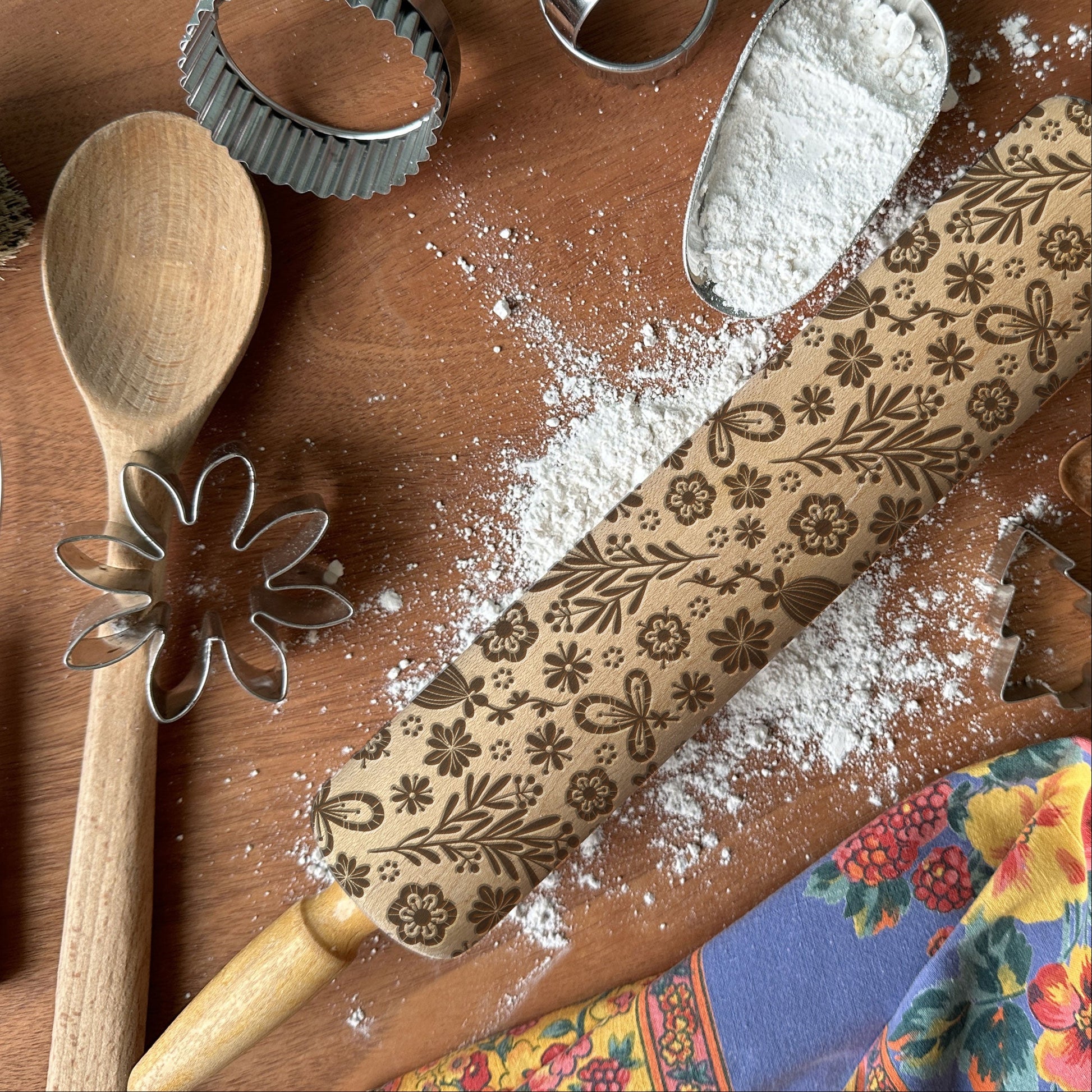 Baking tools including a wooden spoon, rolling pin with floral patterns, and cookie cutters on a wooden surface.