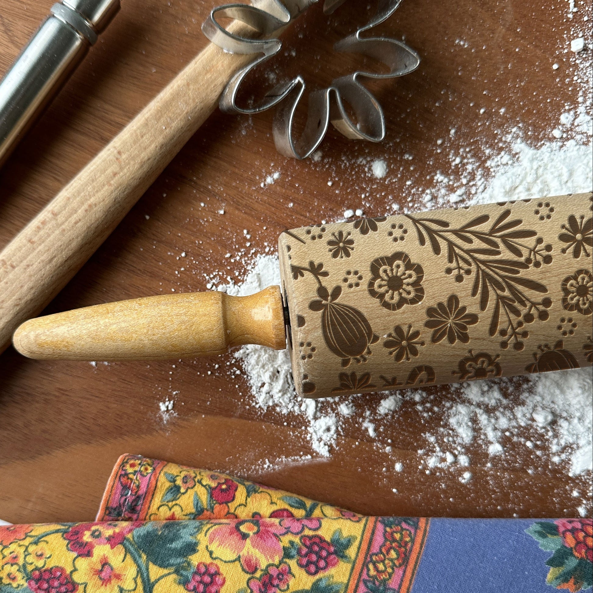 Decorative rolling pin with floral patterns on a wooden surface with flour and cookie cutters.