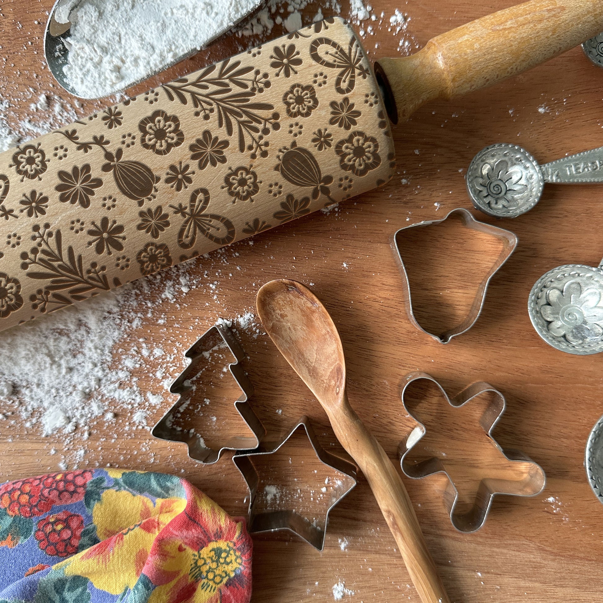 Baking tools including a floral-patterned rolling pin, cookie cutters, and measuring spoons on a wooden surface with flour.