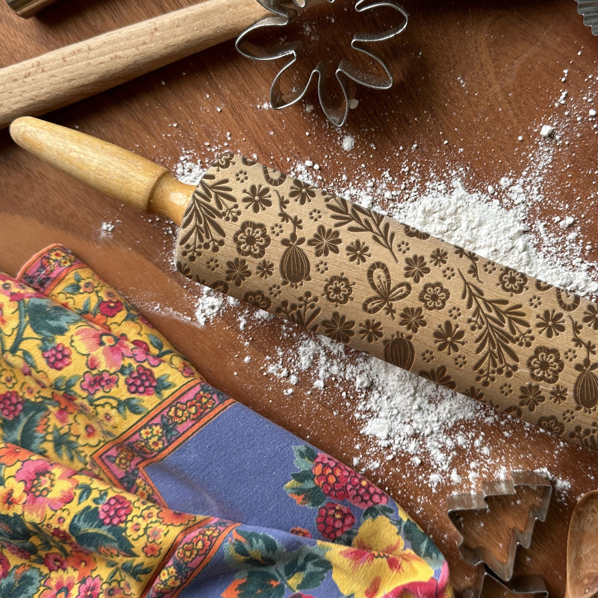 Decorative rolling pin with floral patterns on a wooden surface with flour and cookie cutters.