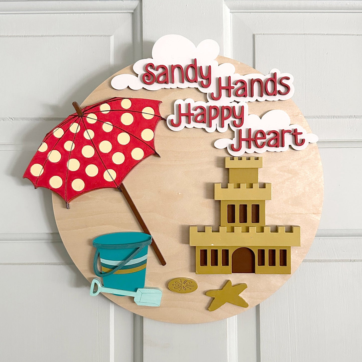 Decorative wall art with sandcastle, umbrella, bucket, and shovel on a wooden circle with 'Sandy Hands Happy Heart' text.