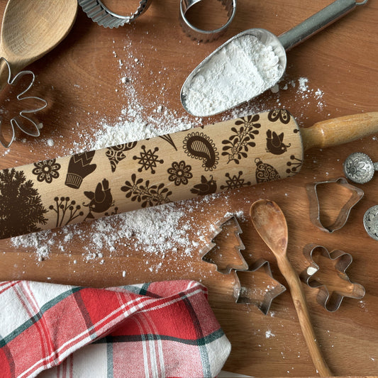 Wooden rolling pin with embossed designs on a wooden surface with flour, cookie cutters, and a plaid cloth.