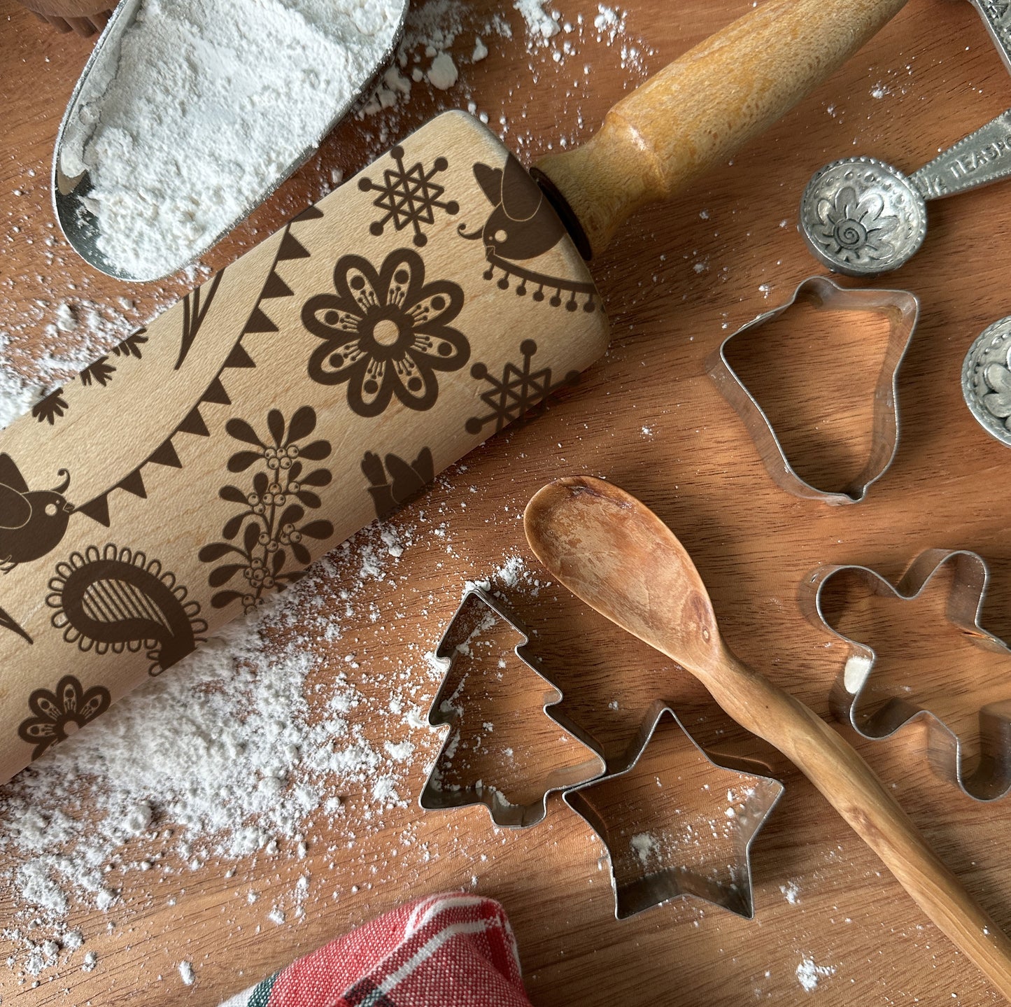 Wooden rolling pin with embossed patterns, flour, metal cookie cutters, and wooden spoon on a wooden surface.