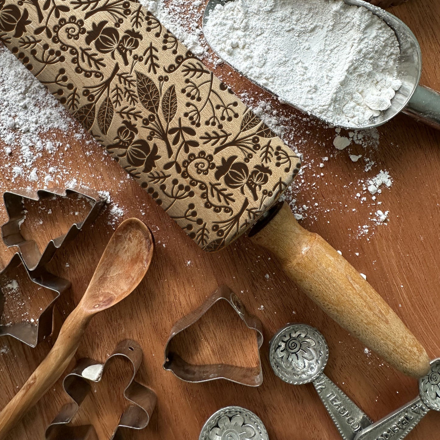 Baking tools including a patterned rolling pin, cookie cutters, and flour on a wooden surface.