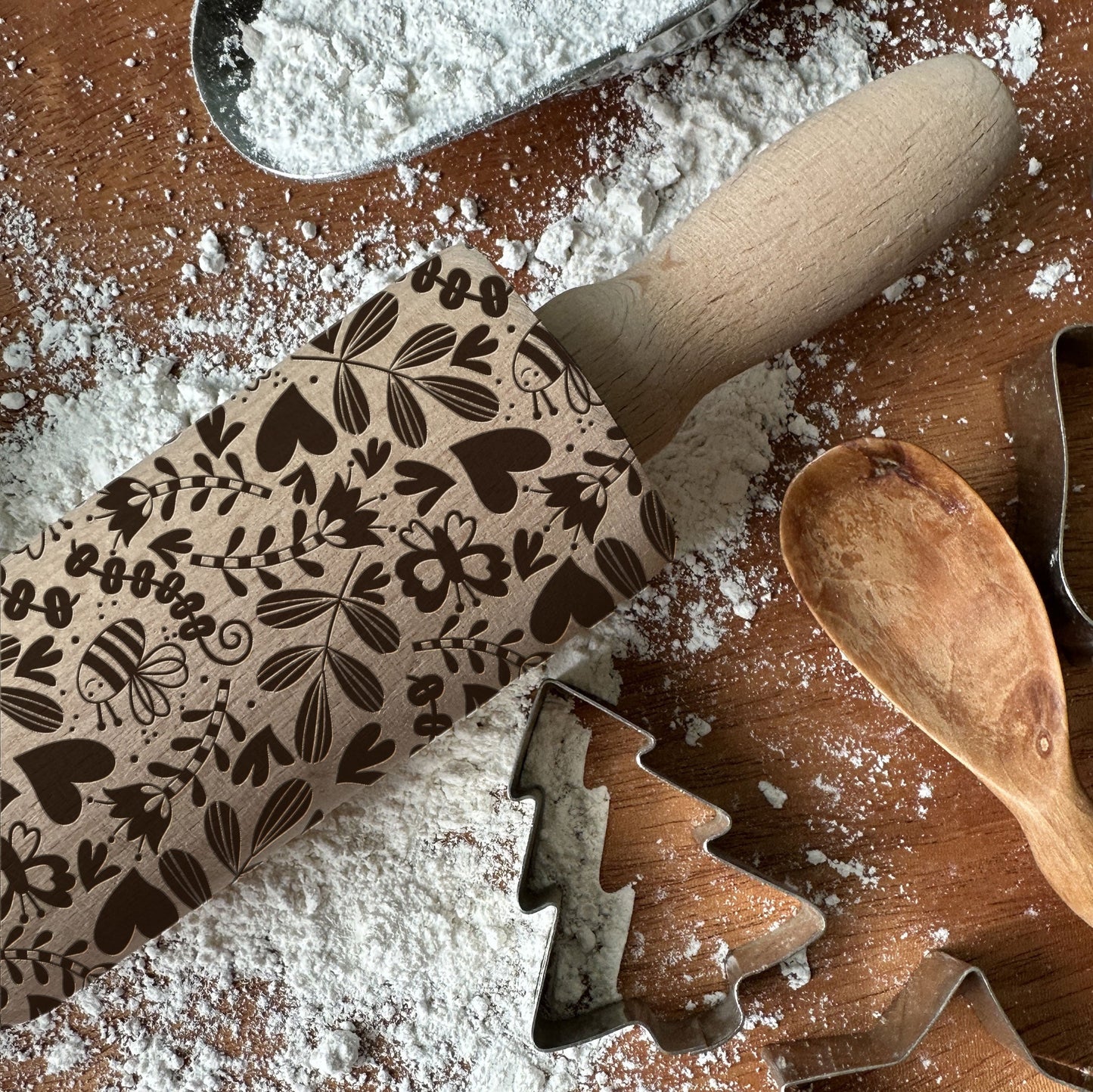 Decorative rolling pin with floral patterns on a wooden surface with flour and cookie cutters.