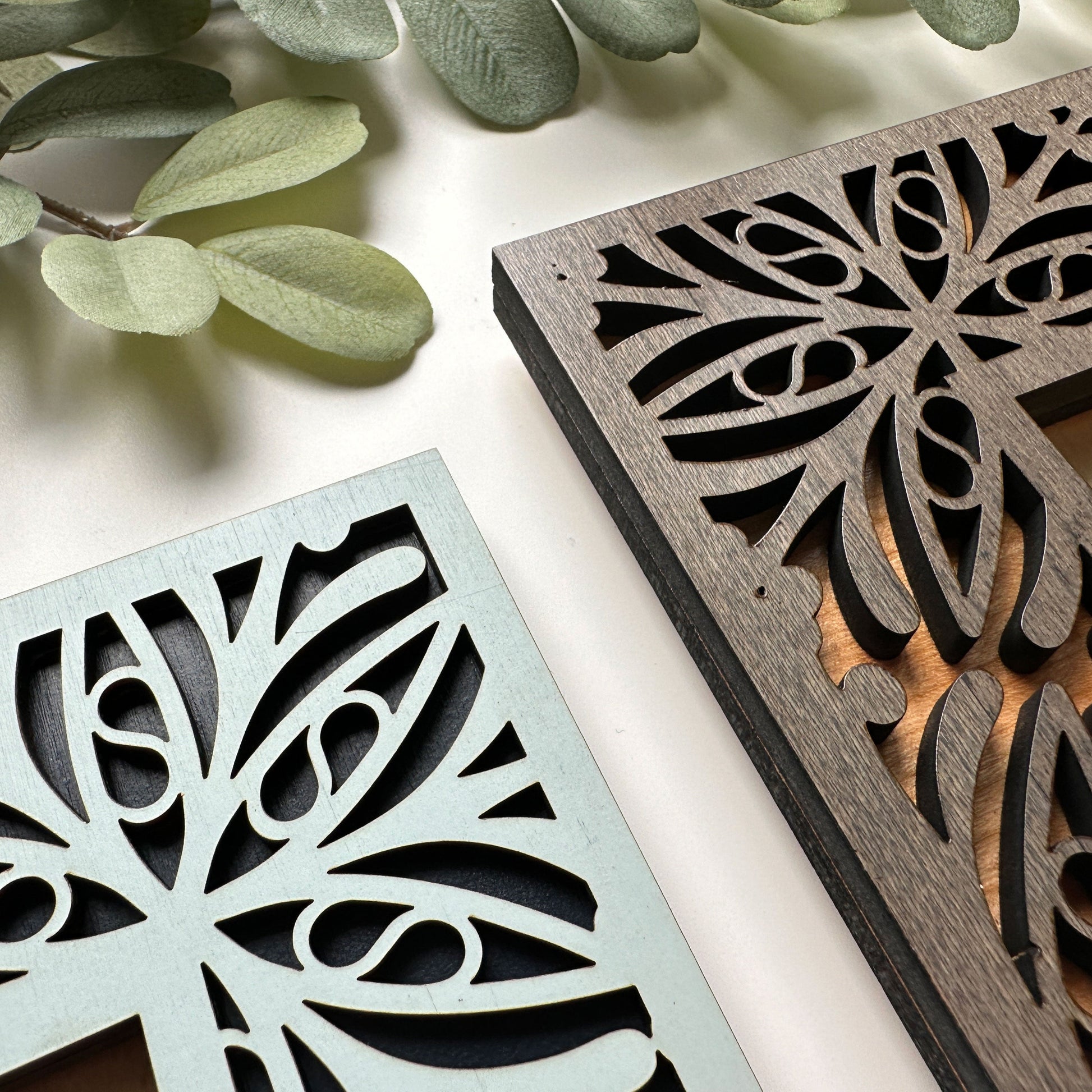 Two decorative metal panels with intricate cut-out patterns on a light surface with green leaves.