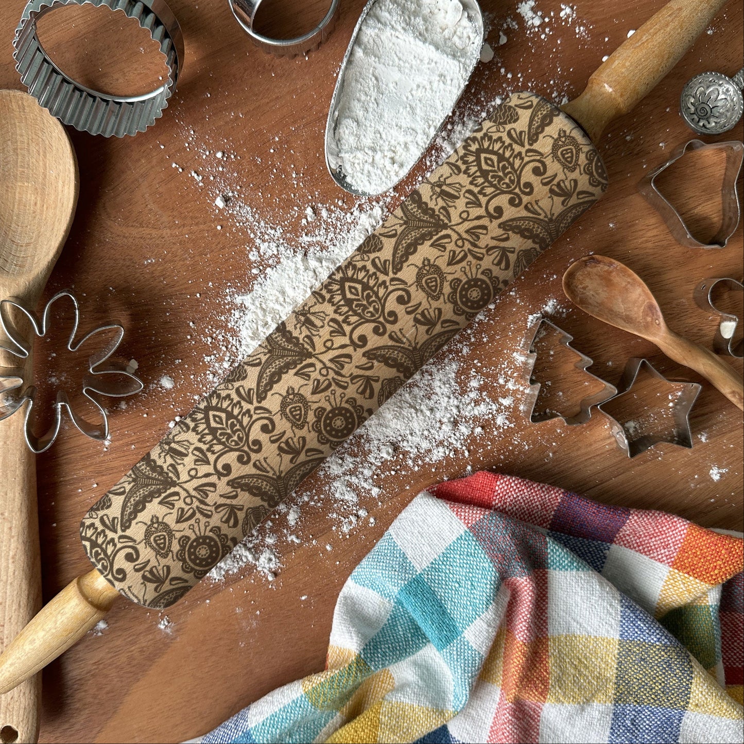Baking tools including a rolling pin with floral patterns, wooden spoon, flour scoop, and cookie cutters on a wooden surface.