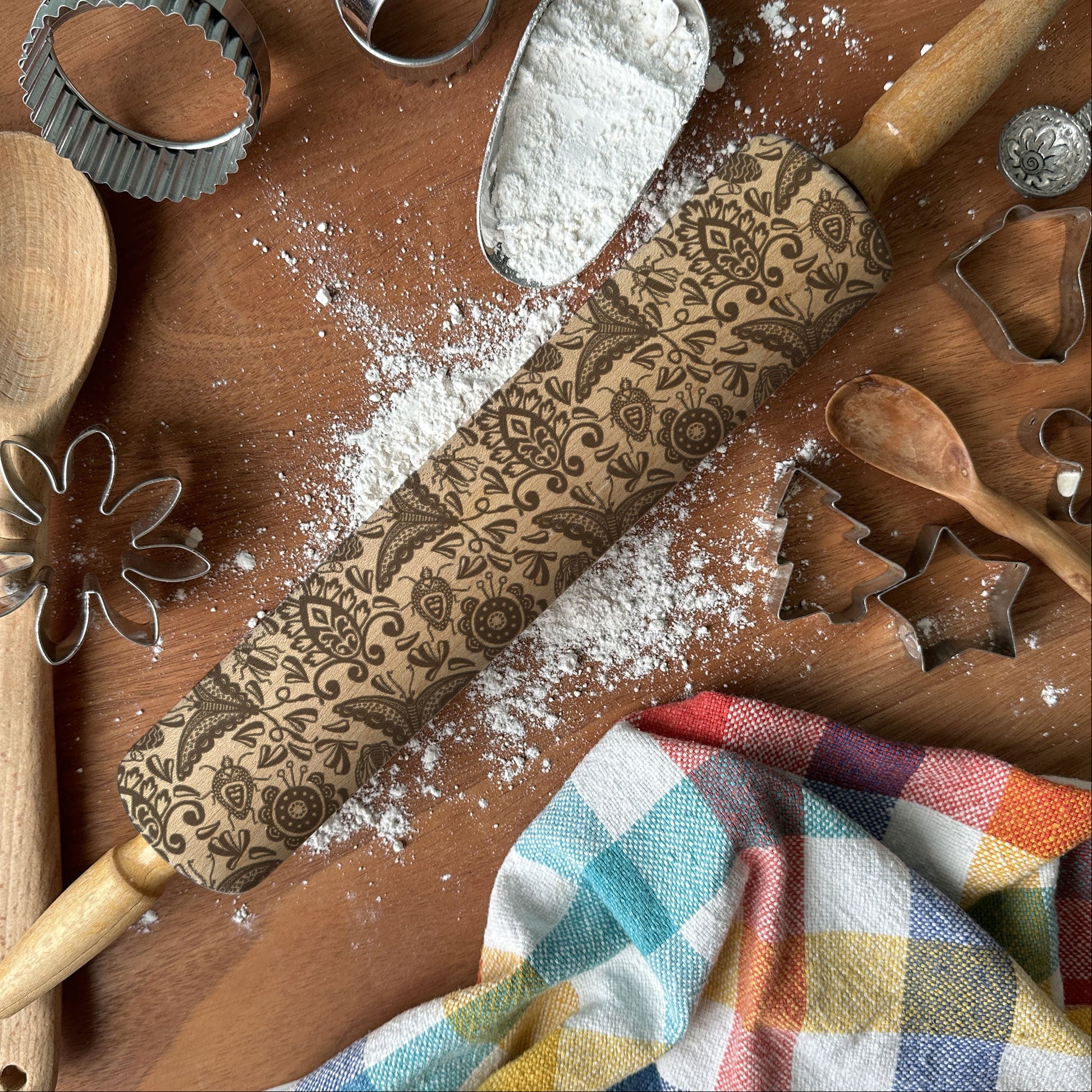 Baking tools including a rolling pin with floral patterns, wooden spoon, flour scoop, and cookie cutters on a wooden surface.
