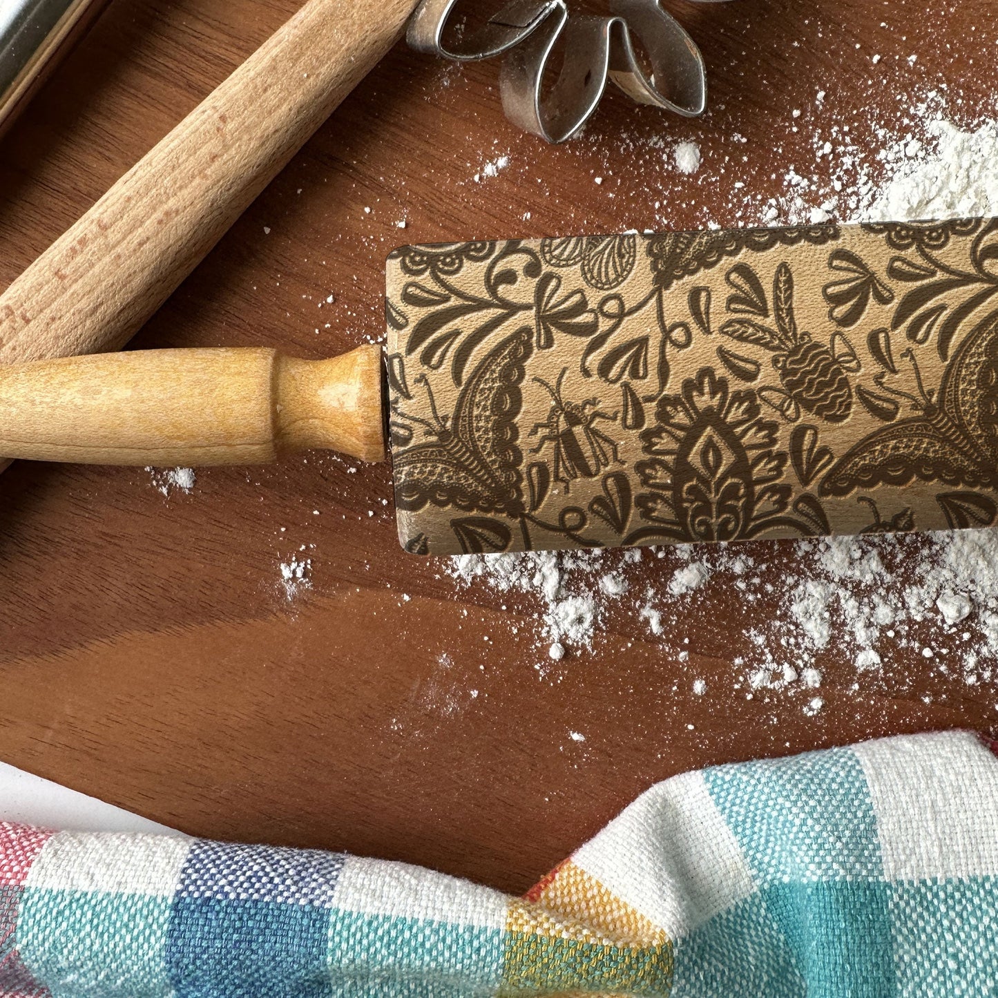 Decorative rolling pin with floral patterns on a wooden surface with flour and a checkered cloth.