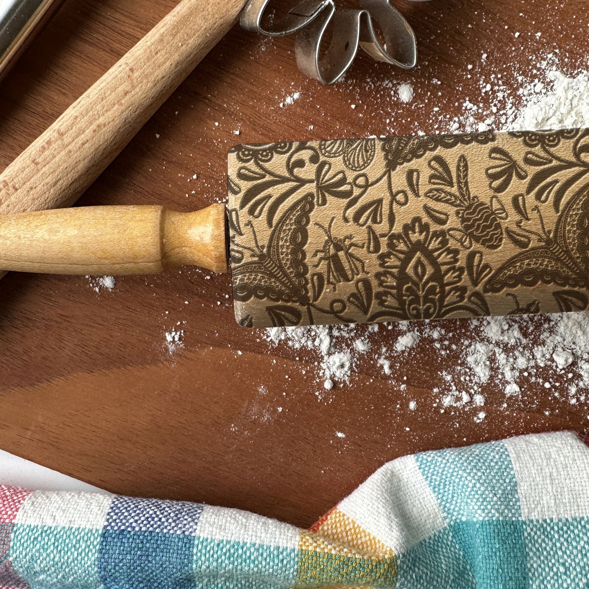 Decorative rolling pin with floral patterns on a wooden surface with flour and a checkered cloth.