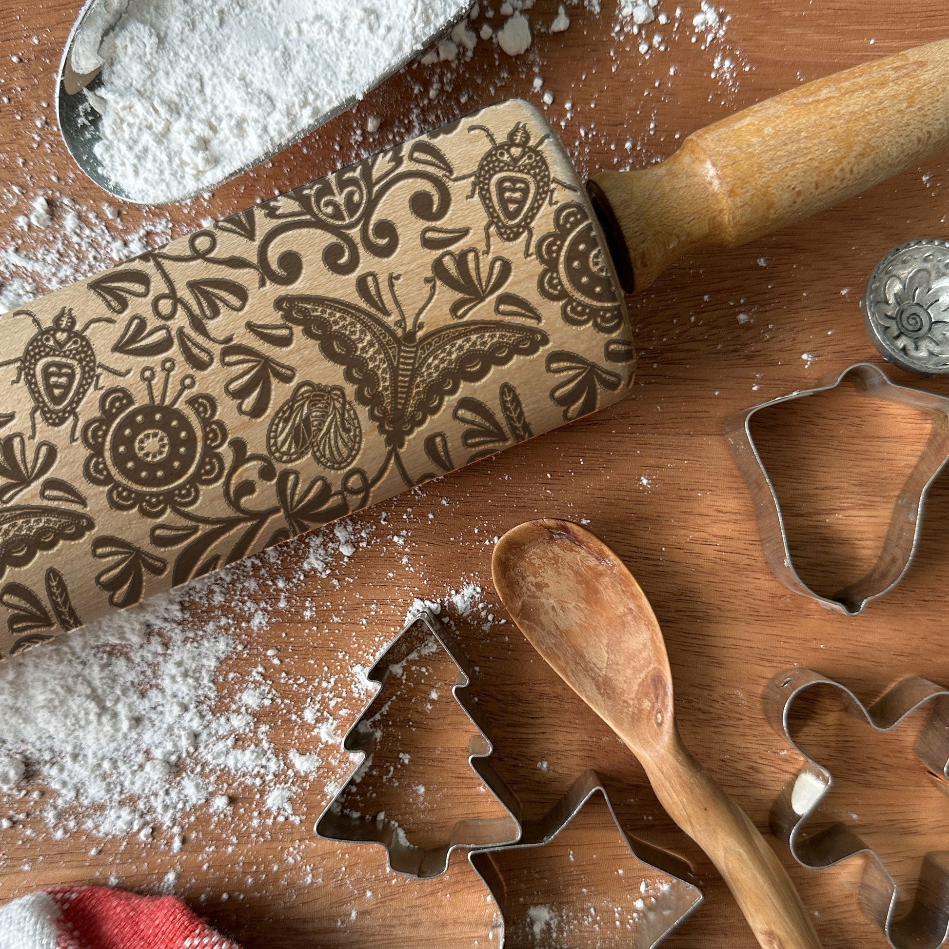 Decorative rolling pin with butterfly design on a wooden surface with cookie cutters and flour.