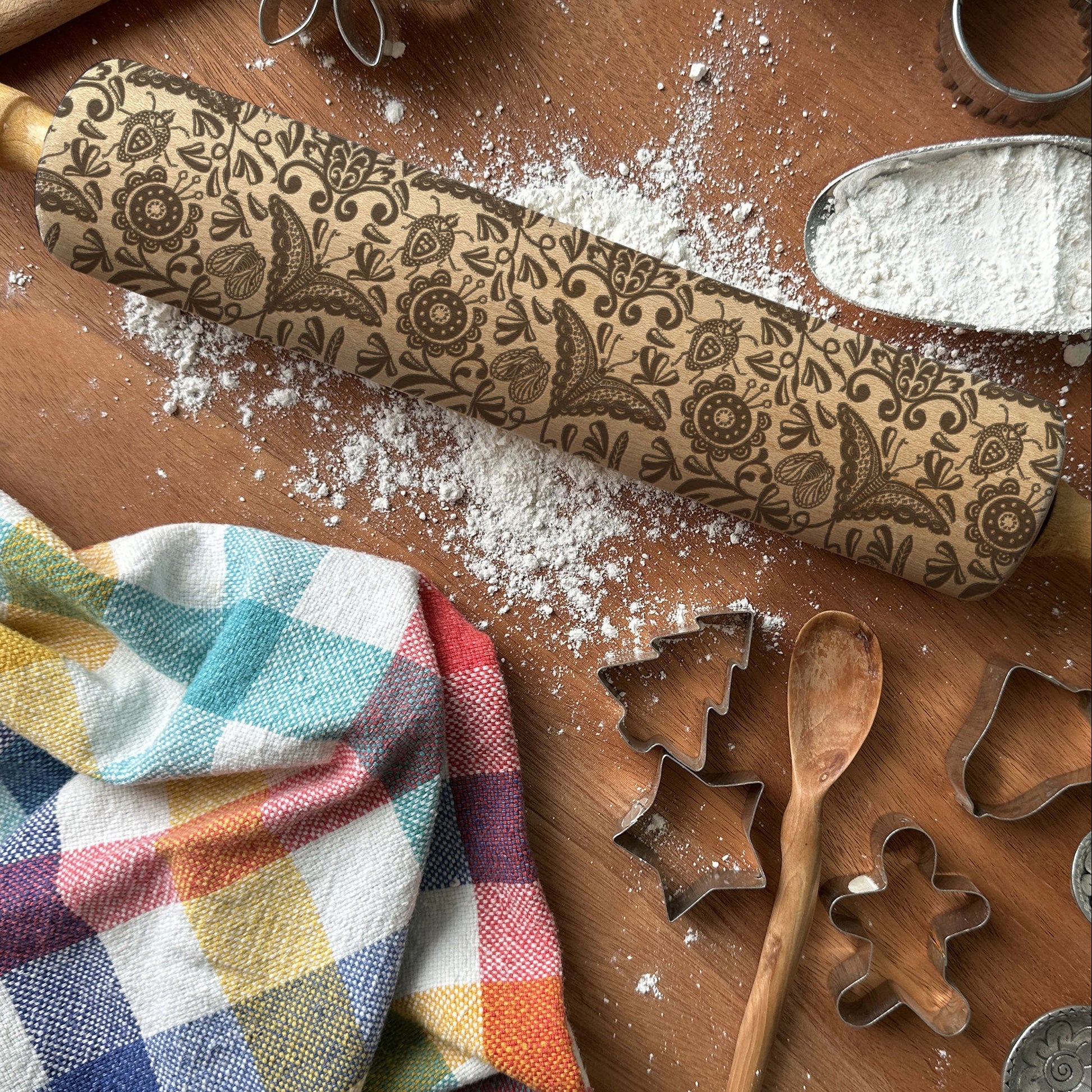 Decorative rolling pin with floral patterns on a wooden surface with flour, cookie cutters, and a colorful towel.