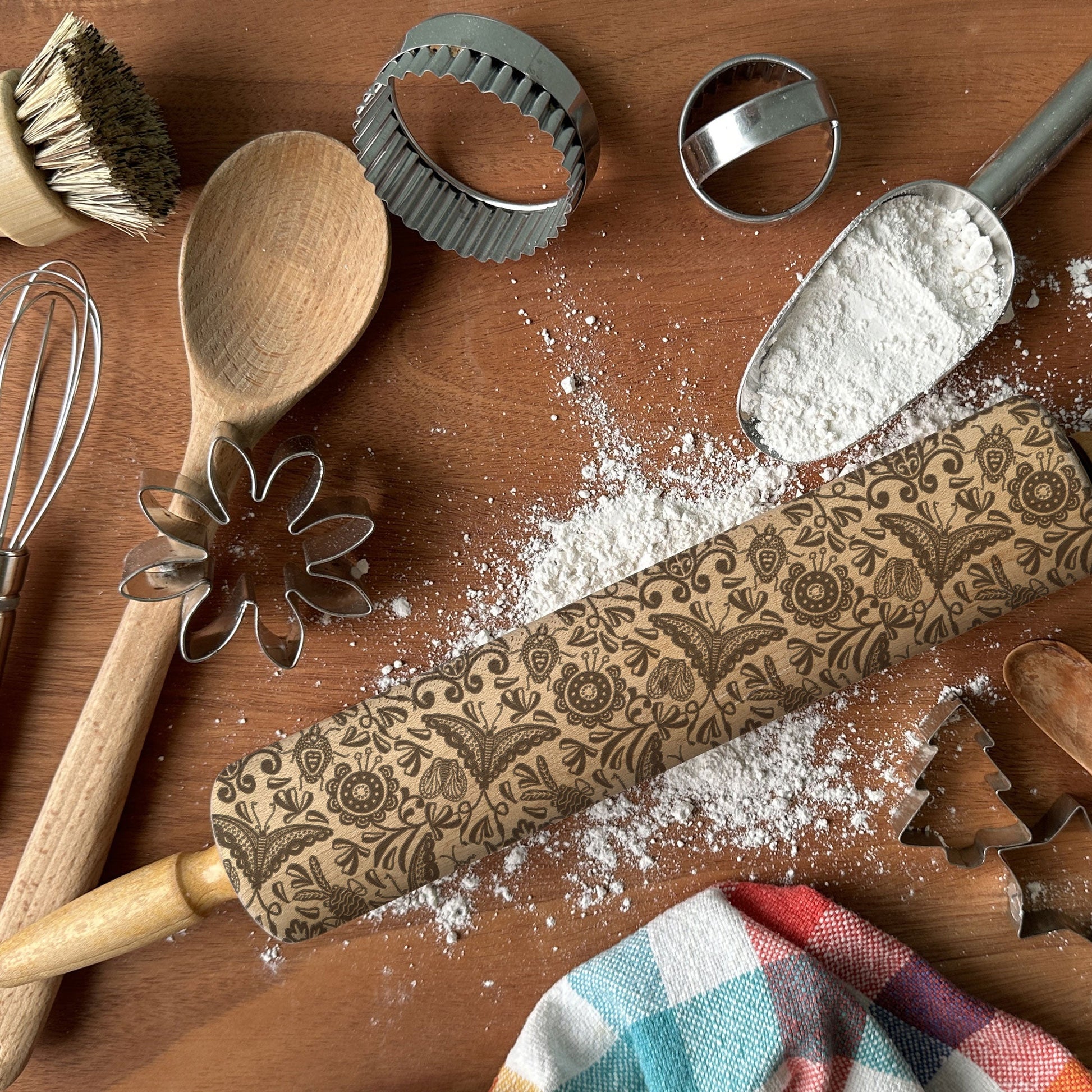 Baking tools including a wooden rolling pin with a decorative pattern, flour, and utensils on a wooden surface.